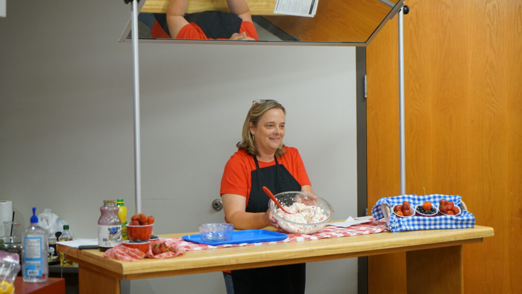 A woman leading a cooking demonstration smiles while holding a bowl of mixed batter