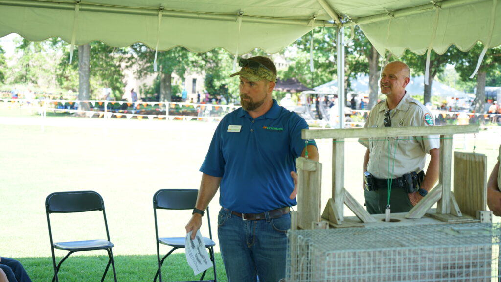 A male speaker gestures to an animal trap during a presentation