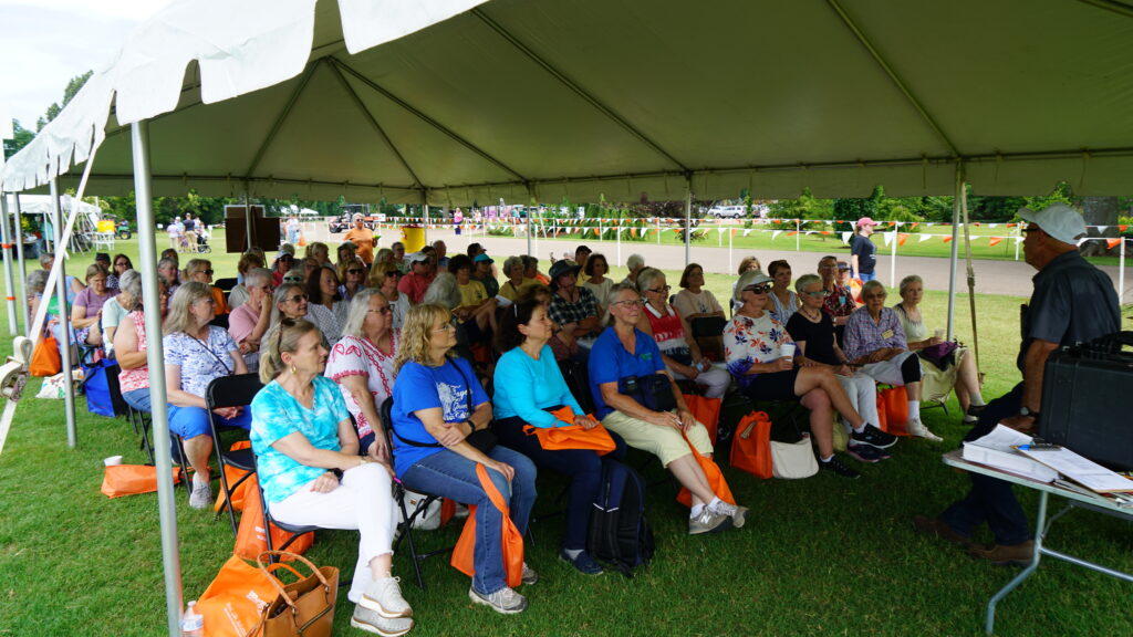 A crowd of people under a large outdoor tent listen to a presenter